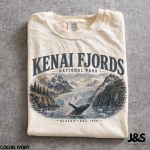 May include: Ivory-colored t-shirt featuring a scenic illustration of Kenai Fjords National Park in Alaska. The design includes mountains, a whale breaching the water, and the text "KENAI FJORDS NATIONAL PARK - ALASKA EST. 1980".