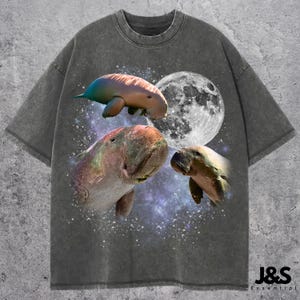 May include: Dark gray t-shirt featuring a graphic design of three manatees, a full moon, and a starry background. The shirt has a crew neck and short sleeves. The brand name "J&S Essential" is printed at the bottom right.