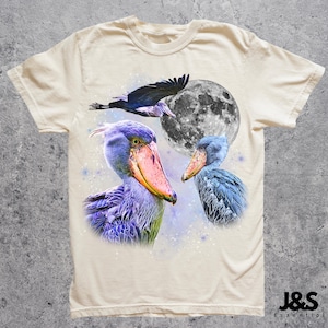 Puede incluir: Camiseta color crema con un dise&ntilde;o gr&aacute;fico de tres aves picozapato, una luna llena y un p&aacute;jaro volando. Las aves est&aacute;n representadas en tonos de azul, morado y rosa. La camiseta tiene cuello redondo y mangas cortas. La marca "J&S Essential" est&aacute; impresa en la parte inferior derecha.