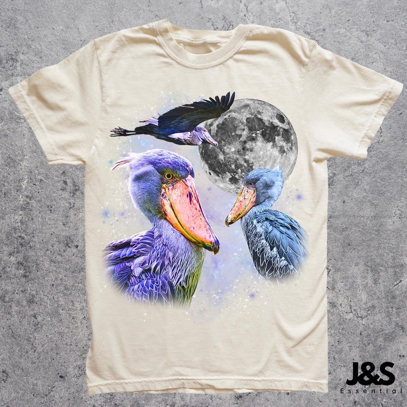 Puede incluir: Camiseta color crema con un dise&ntilde;o gr&aacute;fico de tres aves picozapato, una luna llena y un p&aacute;jaro volando. Las aves est&aacute;n representadas en tonos de azul, morado y rosa. La camiseta tiene cuello redondo y mangas cortas. La marca "J&S Essential" est&aacute; impresa en la parte inferior derecha.