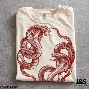 Op de afbeelding: Een ivoorkleurig t-shirt met een gedetailleerde rode afbeelding van twee cobra's die tegenover elkaar staan. De slangen zijn afgebeeld met ingewikkelde schubpatronen en gestileerde wolkenmotieven. Het t-shirt heeft een ronde hals en een label.