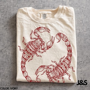 May include: An ivory t-shirt featuring a dark red graphic print of two scorpions. The scorpions are decorated with intricate floral patterns and swirling lines. The shirt has a crew neckline and a Comfort Colors brand label at the inner neck.