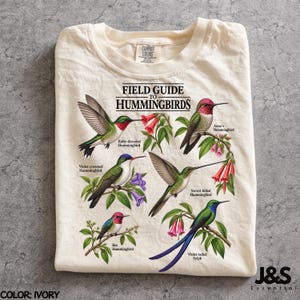May include: An ivory t-shirt featuring a graphic print titled Field Guide to Hummingbirds. The design displays five colourful hummingbird species with their names: Ruby-throated, Anna's, Violet-crowned, Sword-billed, and Violet-tailed Sylph.