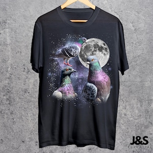 Puede incluir: Camiseta negra con un gr&aacute;fico de tres palomas con plumas iridiscentes sobre un cielo estrellado y una luna llena. La camiseta tiene el texto "J&S Essential" en la parte inferior.
