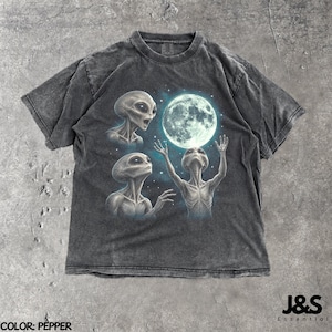 May include: A pepper-coloured, short-sleeve t-shirt featuring a graphic print of three grey extraterrestrial figures reaching towards a large, glowing full moon surrounded by small white stars against a dark blue nebula background.