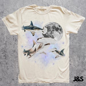 Puede incluir: Camiseta color crema con un dise&ntilde;o de tiburones nadando alrededor de una luna llena. Los tiburones son de varios tonos de gris y blanco, con un fondo estrellado. La camiseta tiene cuello redondo y mangas cortas.