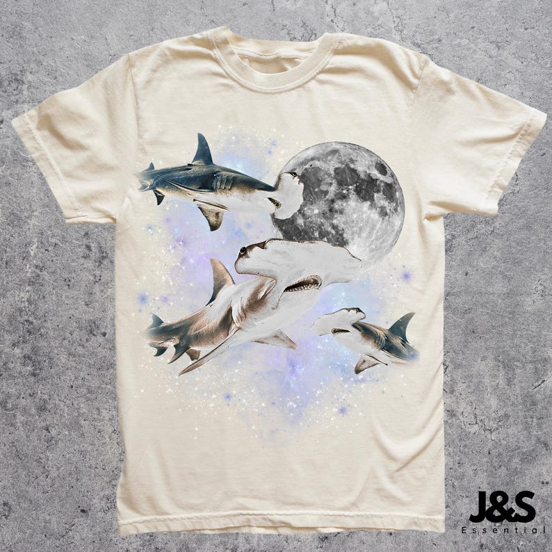 Puede incluir: Camiseta color crema con un dise&ntilde;o de tiburones nadando alrededor de una luna llena. Los tiburones son de varios tonos de gris y blanco, con un fondo estrellado. La camiseta tiene cuello redondo y mangas cortas.
