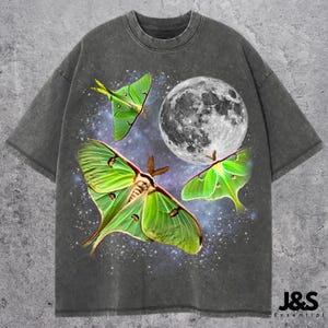 May include: Dark gray t-shirt featuring a graphic of three green Luna moths, a full moon, and a starry background. The shirt has a vintage, washed-out look. The text "J&S Essential" is at the bottom right.