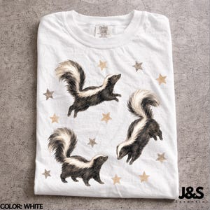 Puede incluir: Camiseta blanca con un diseño de acuarela de tres zorrillos con pelaje blanco y negro y colas esponjosas. Estrellas color canela están dispersas alrededor de los zorrillos. La camiseta tiene cuello redondo y el texto "J&S Essential" es visible.