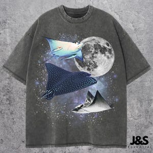 Puede incluir: Camiseta gris oscuro con un diseño celestial. Presenta tres rayas, una luna llena y un fondo estrellado. La camiseta tiene un efecto de lavado vintage y el texto "J&S Essential" es visible en la parte inferior derecha.