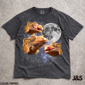 Könnte beinhalten: Ein anthrazitfarbenes Kurzarm-T-Shirt mit einem Grafikdruck von drei Kronengeckos vor dem Hintergrund eines Vollmonds und einer Sternengalaxie. Die Geckos sind in Orange- und Brauntönen mit sichtbaren Augen und Zungen dargestellt.
