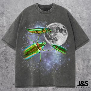 Puede incluir: Camiseta gris oscuro con un gráfico de tres escarabajos iridiscentes, una luna llena y una galaxia estrellada. La camiseta tiene cuello redondo y mangas cortas. El logo de la marca "J&S Essential" está en la parte inferior derecha.