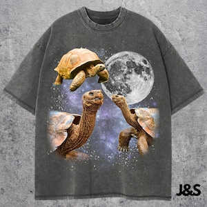 Pode incluir: Camiseta cinza escuro com um gráfico de três tartarugas, uma lua cheia e um fundo de galáxia estrelada. A camiseta tem gola redonda e mangas curtas. O texto "J&S Essential" está impresso no canto inferior direito.