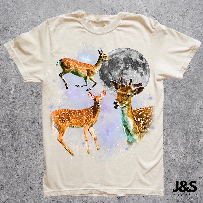May include: Cream-colored t-shirt featuring a design with three deer, a full moon, and a starry background. The deer are brown with white spots. The text "J&S Essential" is at the bottom right.