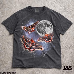 May include: A charcoal gray short-sleeve t-shirt featuring a graphic print of three large moths with orange and black wings flying against a backdrop of a full moon and a starry night sky. The shirt has a crew neckline and a faded, washed-out texture.