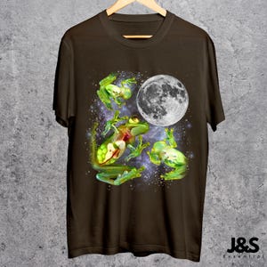 May include: A brown t-shirt featuring a whimsical design of green frogs and a full moon against a starry background. The frogs appear to be floating in space. The shirt is hanging on a wooden hanger.