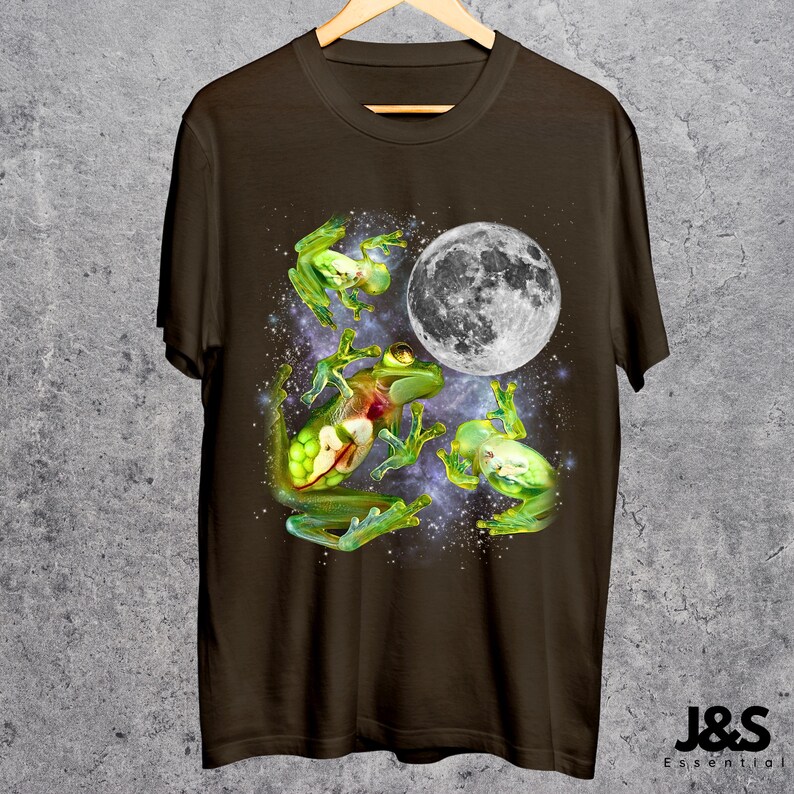 May include: A brown t-shirt featuring a whimsical design of green frogs and a full moon against a starry background. The frogs appear to be floating in space. The shirt is hanging on a wooden hanger.