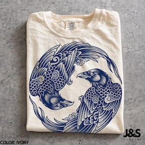 May include: An ivory t-shirt featuring a circular graphic of two dark blue ravens arranged in a yin-yang formation. The birds are depicted with detailed feathers and stylised cloud patterns. The shirt has a crew neckline and a Comfort Colors brand label.
