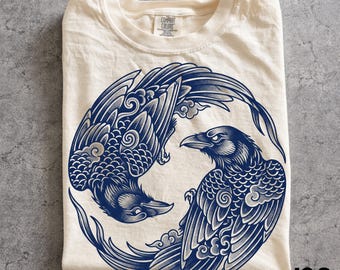 May include: An ivory t-shirt featuring a circular graphic of two dark blue ravens arranged in a yin-yang formation. The birds are depicted with detailed feathers and stylized cloud patterns. The shirt has a crew neckline and a Comfort Colors brand label.