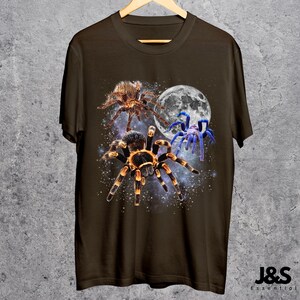 May include: A brown t-shirt featuring a graphic design of three spiders against a starry background and a full moon. The spiders are in shades of brown, orange, and blue. The shirt is hanging on a wooden hanger.