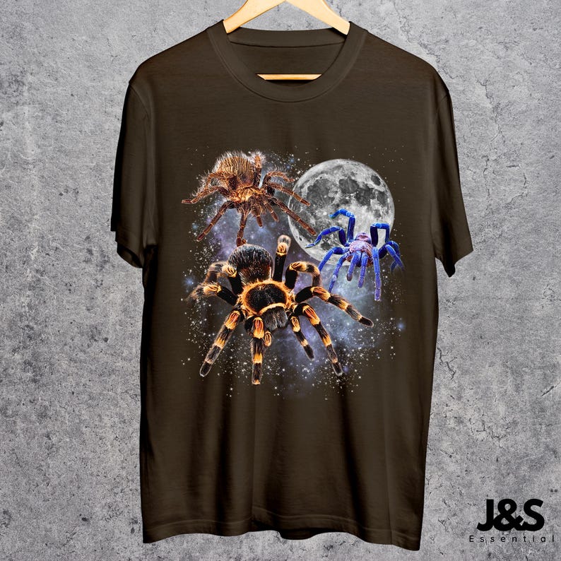 May include: A brown t-shirt featuring a graphic design of three spiders against a starry background and a full moon. The spiders are in shades of brown, orange, and blue. The shirt is hanging on a wooden hanger.