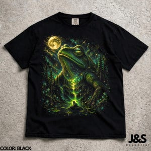 Può includere: T-shirt nera con un'illustrazione dettagliata di una rana che guarda la luna piena. Il design include una foresta, una cascata e un cielo stellato, con accenti verdi e gialli. Il testo "J&S Essential" è in basso.