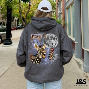 May include: Dark gray hoodie with a whimsical zebra design. The back features three zebras, a full moon, and a starry background. The text "J&S Essential" is at the bottom right.