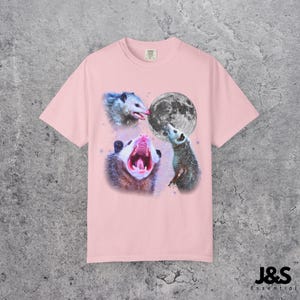 Pode incluir: Camiseta rosa claro com um design gr&aacute;fico de tr&ecirc;s gamb&aacute;s e uma lua cheia. Os gamb&aacute;s est&atilde;o em v&aacute;rias poses, um com a boca aberta. O texto "J&S Essential" est&aacute; no canto inferior direito.