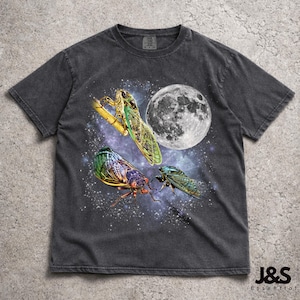 May include: Dark gray t-shirt featuring a graphic of three cicadas, a full moon, and a starry night sky. The cicadas are in shades of green, purple, and blue. The shirt has a vintage, worn look.