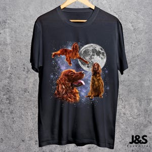 K&ouml;nnte beinhalten: Schwarzes T-Shirt mit einem Design von drei Irish Setter Hunden vor einem Sternenhimmel und einem Vollmond. Die Hunde sind in verschiedenen Posen dargestellt, darunter einer in der Luft, einer sitzend und einer mit herausgestreckter Zunge.
