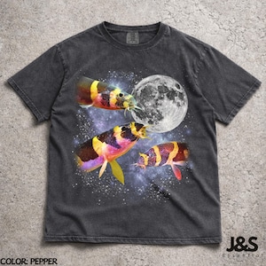 Puede incluir: Camiseta gris oscuro con un diseño gráfico de peces coloridos en el espacio, con una luna llena. Los peces tienen detalles amarillos, naranjas y morados. La camiseta tiene el texto "J&S Essential" en la parte inferior. Color: pimienta.