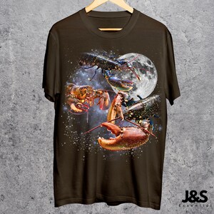 以下が含まれることがあります： 宇宙にいるロブスターのグラフィックが描かれた濃い茶色のTシャツ。満月と星空を背景に、赤、オレンジ、黒の色合いでロブスターが描かれています。Tシャツは木製のハンガーに掛けられています。