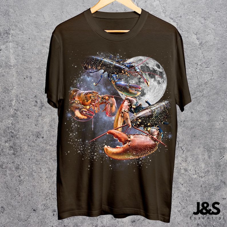 以下が含まれることがあります： 宇宙にいるロブスターのグラフィックが描かれた濃い茶色のTシャツ。満月と星空を背景に、赤、オレンジ、黒の色合いでロブスターが描かれています。Tシャツは木製のハンガーに掛けられています。