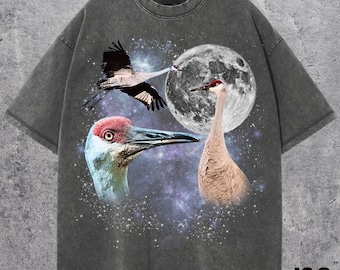 May include: Dark gray t-shirt featuring a graphic of three cranes, a full moon, and a starry sky. The cranes are depicted in flight and in profile, with red and white accents. The shirt has a vintage, washed look, and the text "J&S Essential" is printed at the bottom.