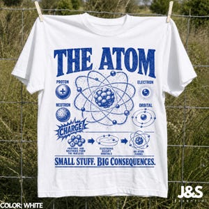 May include: A white short-sleeve t-shirt featuring a blue graphic print titled THE ATOM. The design includes scientific diagrams of protons, neutrons, electrons, and orbitals with the text SMALL STUFF. BIG CONSEQUENCES. and CHARGE!