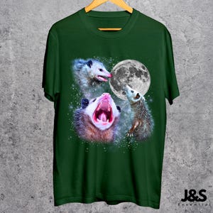 Pode incluir: T-shirt verde escuro com um desenho gr&aacute;fico de tr&ecirc;s gamb&aacute;s e uma lua cheia. Um gamb&aacute; boceja. O design inclui um fundo estrelado. A t-shirt est&aacute; pendurada num cabide de madeira.