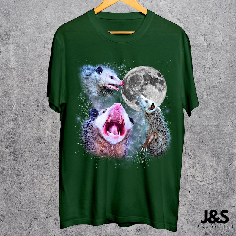 Pode incluir: T-shirt verde escuro com um desenho gr&aacute;fico de tr&ecirc;s gamb&aacute;s e uma lua cheia. Um gamb&aacute; boceja. O design inclui um fundo estrelado. A t-shirt est&aacute; pendurada num cabide de madeira.