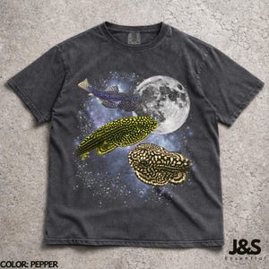 May include: Pepper-colored t-shirt featuring a graphic design of fish in space with a full moon. The design includes three fish with unique patterns against a starry background. The text "J&S Essential" is in the lower right corner.