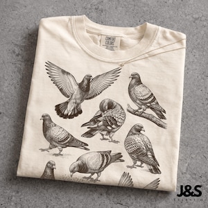 May include: Cream-colored t-shirt featuring a detailed illustration of several pigeons in various poses. The design includes a pigeon with outstretched wings, others perched or preening. The shirt has a crew neck and a tag that reads "COMFORT COLORS". The brand name "J&S Essential" is printed on the bottom right.