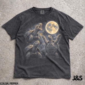 Puede incluir: Camiseta color pimienta con un diseño caprichoso de tres tortugas bajo una luna llena. Una tortuga bosteza, otra tiene los brazos levantados y la tercera mira hacia arriba. La camiseta tiene un aspecto vintage y desgastado.