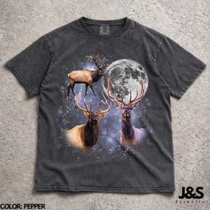 May include: Pepper-colored t-shirt featuring a graphic of three elk with large antlers, a full moon, and a starry background. The text "J&S Essential" is in the lower right corner. The shirt is a crew neck.