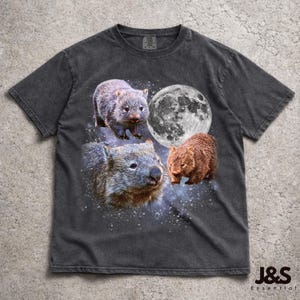 Puede incluir: Camiseta gris oscuro con un diseño de wombat. El gráfico incluye tres wombats, una luna llena y un fondo estrellado. La camiseta tiene un aspecto vintage y desgastado, con el texto "J&S Essential" impreso en la parte inferior derecha.