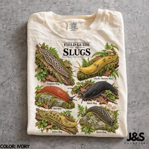 Könnte beinhalten: Ein elfenbeinfarbenes T-Shirt mit einem Grafikdruck mit dem Titel Field Guide to Slugs. Das Design zeigt sechs illustrierte Nacktschnecken, beschriftet als Leopard Slug, Pacific Banana Slug, Red Slug, Black Slug, Yellow Slug und Kerry Slug auf Moos.