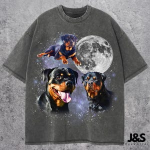 Może przedstawiać: Ciemnoszara koszulka z grafiką przedstawiającą trzy psy rasy Rottweiler i pełnię księżyca na tle gwiezdnego nieba. Koszulka ma wygląd vintage. Tekst "J&S Essential" jest wydrukowany w prawym dolnym rogu.