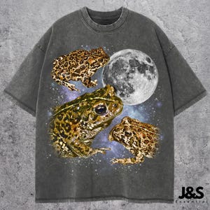 May include: Dark gray t-shirt featuring a graphic of three frogs and a full moon against a starry background. The frogs have brown and green patterns. The shirt has a vintage, washed look.