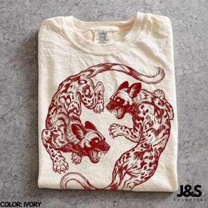 May include: An ivory t-shirt featuring a dark red graphic illustration of two snarling African wild dogs arranged in a circular, yin-yang style composition. The shirt has a crew neckline and a Comfort Colors brand label at the inner collar.