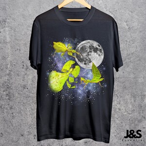 May include: Black t-shirt featuring a vibrant graphic of leaf insects, a full moon, and a starry galaxy background. The design includes detailed illustrations of the insects in shades of green and yellow. The shirt is hanging on a wooden hanger.