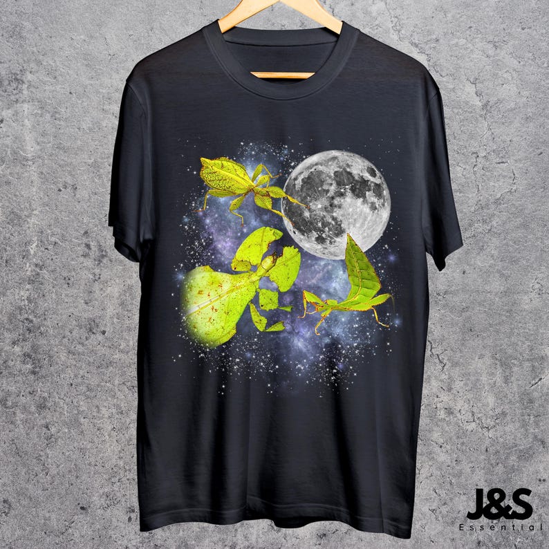 May include: Black t-shirt featuring a vibrant graphic of leaf insects, a full moon, and a starry galaxy background. The design includes detailed illustrations of the insects in shades of green and yellow. The shirt is hanging on a wooden hanger.