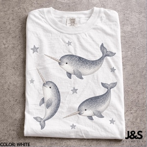 Peut inclure: T-shirt blanc avec trois narvals aquarelles et plusieurs étoiles grises. Les narvals ont des corps gris et de longues défenses blanches. Le t-shirt est de Comfort Colors et porte le logo J&S Essential.