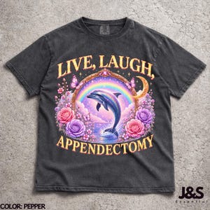 May include: Pepper-colored t-shirt featuring a whimsical design. The graphic includes a dolphin leaping through a rainbow, surrounded by flowers and butterflies, with the text "LIVE, LAUGH, APPENDECTOMY".
